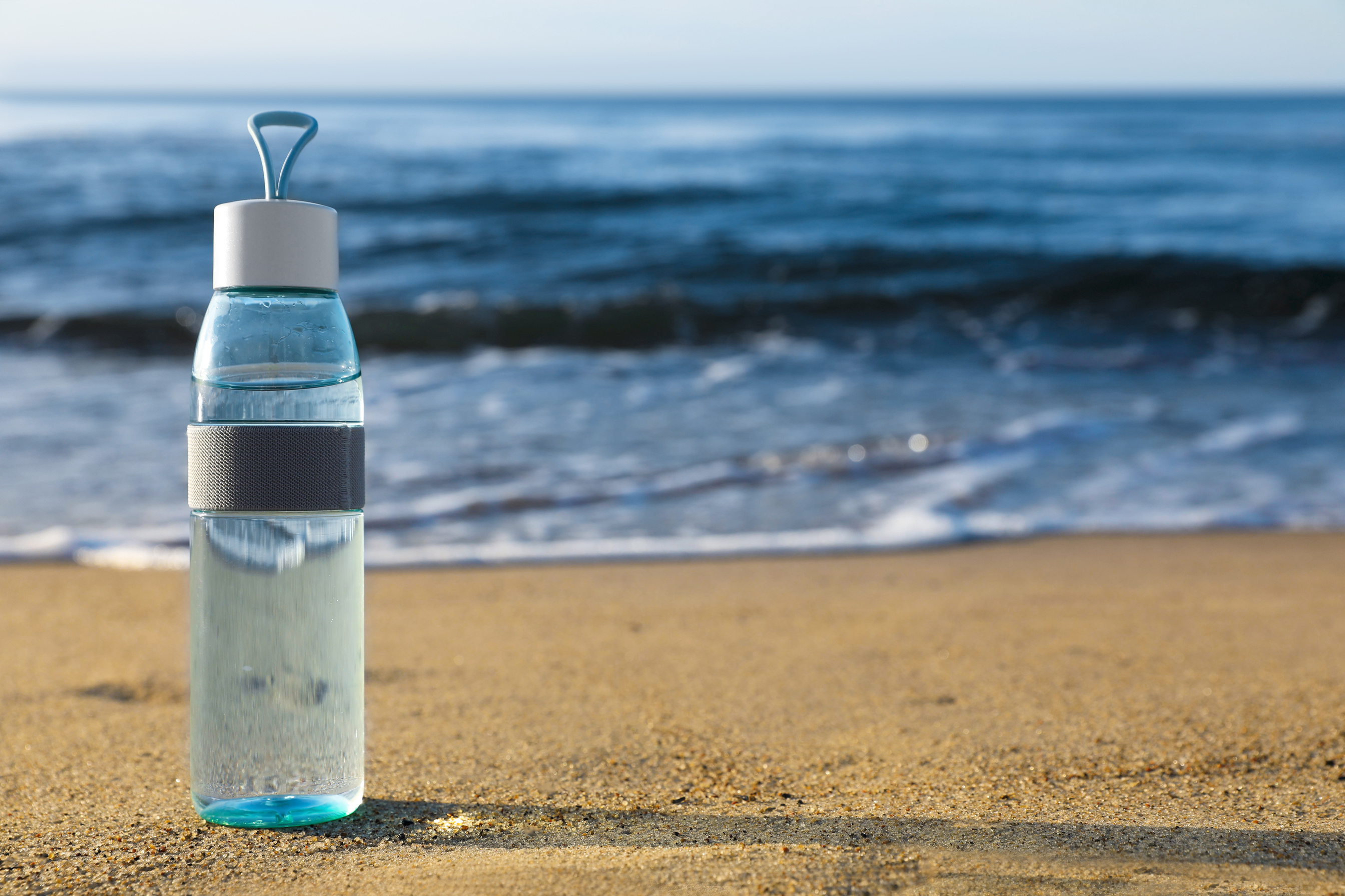 Glass Bottle with Water on Wet Sand near Sea. Space for Text